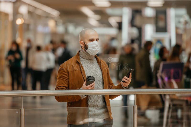 Mann in einer Gesichtsmaske benutzt ein Telefon und hält einen Kaffee im Einkaufszentrum lizenzfreie stockfotografie