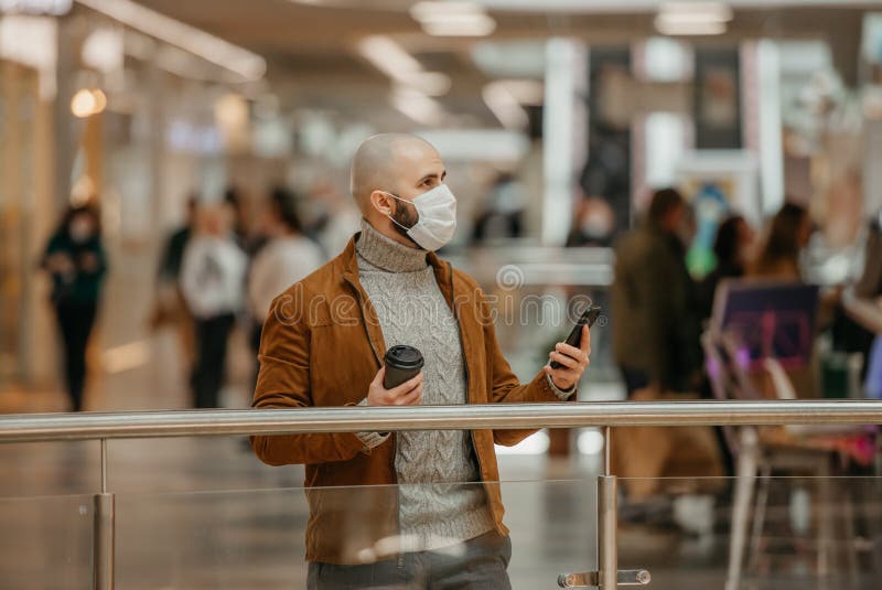 Mann in einer Gesichtsmaske benutzt ein Telefon und hält einen Kaffee im Einkaufszentrum stockfotos