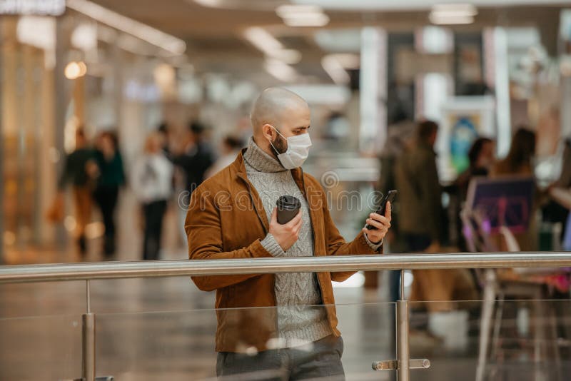 Mann in einer Gesichtsmaske benutzt ein Telefon und hält einen Kaffee im Einkaufszentrum stockbild