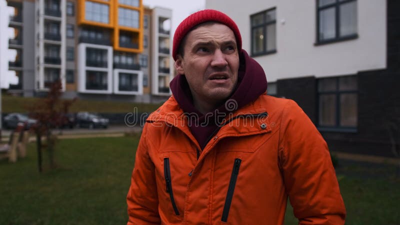 Mann in einer orangefarbenen Jacke mit einem roten Hut bläst sich die Nase im Freien an einem kalten Tag in der Nähe moderner Gebä stock footage
