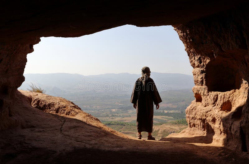 MANN MIT DJELLABA UND PALÄSTINENSISCHEM SCHAL, DIE ZUM TAL VON EINER HOHEN HÖHLE SCHAUEN stockfoto