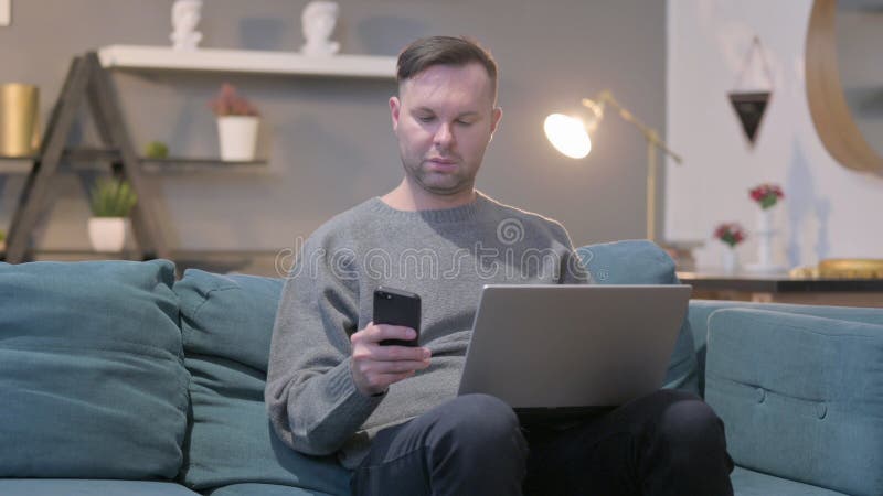 Mann mit Laptop mit Smartphone auf Sofa stockfotografie