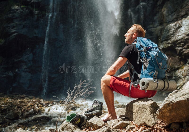 Mann mit Rucksack in aktiven Trekking-Kleidung gekleidet nimmt Trekking Stiefel sitzen in der Nähe von Bergfluss Wasserfall und ge lizenzfreies stockfoto