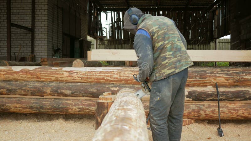 Mann schneidet hölzerne Kettensäge für zukünftiges Haus Schützende Gesichtsmaske auf dem Gesicht des Erbauers und vielen Sägemehl