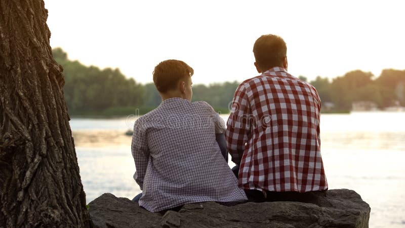 Mannelijke tiener en zijn vaderzitting op steen dichtbij meer en het spreken over het leven royalty-vrije stock foto Mannelijke tiener en zijn vaderzitting op steen dichtbij meer en het spreken over het leven royalty-vrije stock foto
