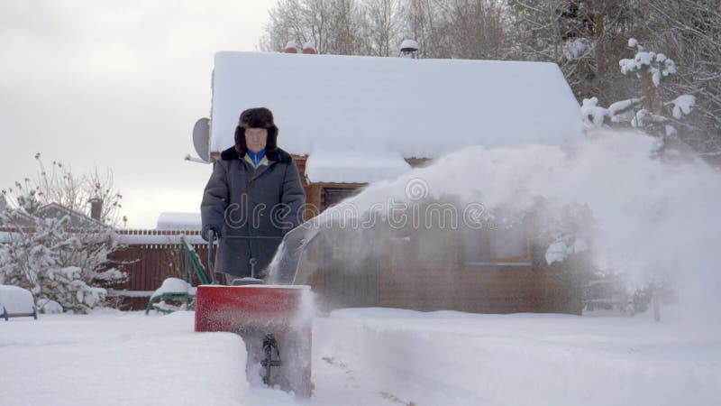 Mannen gör ren snö med bakgrund för snöplogen av trähuset i vinter