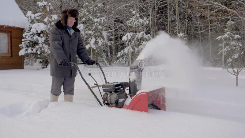 Mannen gör ren snö med bakgrund för snöplogen av trähuset i vinter