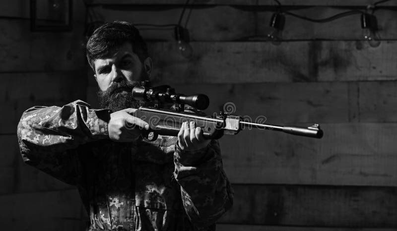 Mannen med skägget bär kamouflagekläder, träinre bakgrund Jägare brutal hipster med vapnet som för siktar royaltyfri bild