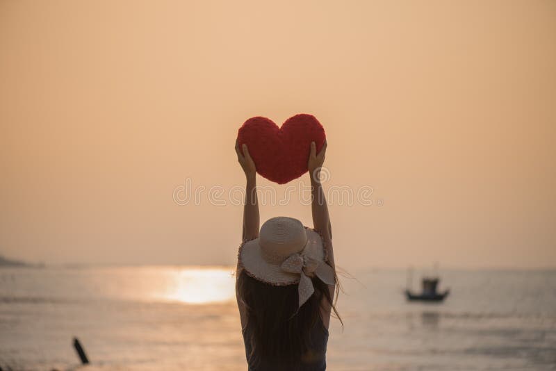 Woman hand holding a red pillow in heart shaped on the beach during sunset for Valentines day and love concept. Woman hand holding a red pillow in heart shaped on the beach during sunset for Valentines day and love concept.