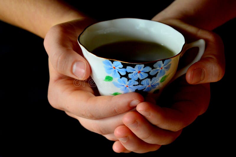 A pair of hands holding a cup of tea in a cute tea cup with blue flowers on black background. A winter time comfort. A pair of hands holding a cup of tea in a cute tea cup with blue flowers on black background. A winter time comfort.