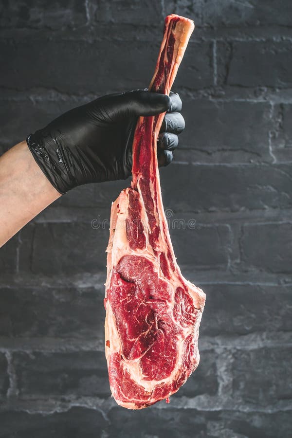 Manos masculinas del filete de carne de vaca de la hacha de guerra de la tenencia del carnicero o del cocinero en fondo rústico o imágenes de archivo libres de regalías Manos masculinas del filete de carne de vaca de la hacha de guerra de la tenencia del carnicero o del cocinero en fondo rústico o imágenes de archivo libres de regalías