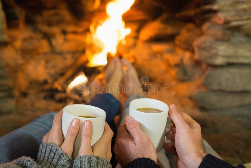 Manos que sostienen las tazas de café delante de la chimenea encendida imagenes de archivo Manos que sostienen las tazas de café delante de la chimenea encendida imagenes de archivo