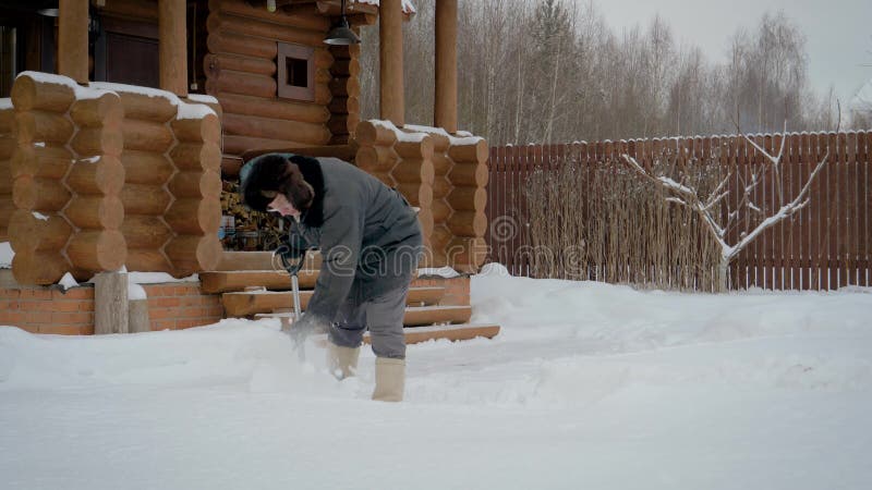 Manrengöringar skyfflar i vintergård från snö på bakgrunden av ett trähus