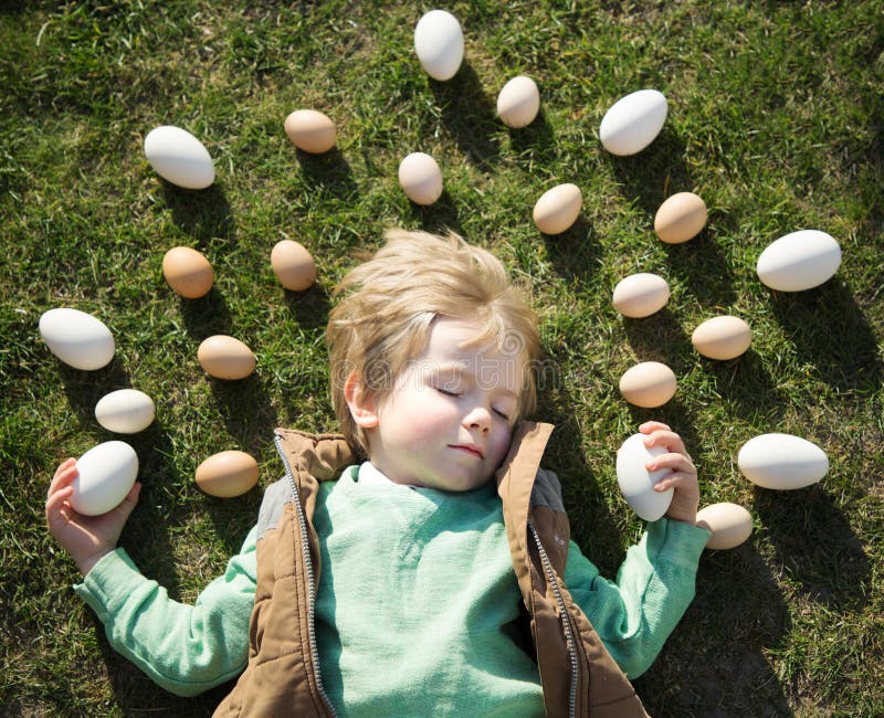 Many white and beige chicken and goose eggs are laid out around the head of a 4-year-old blond boy sleeping on grass