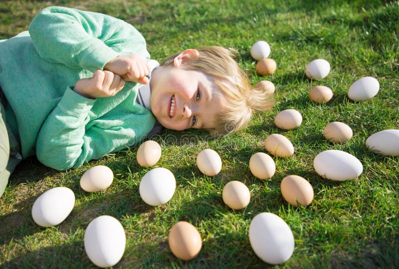 Many white and beige chicken and goose eggs lie on green grass around head of a cheerful boy