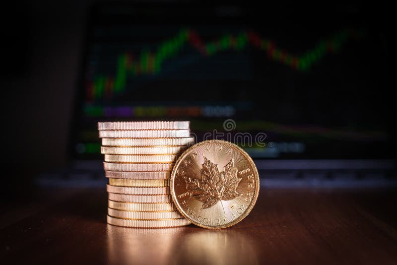 A Maple leaf gold coin stands in front of a stack of gold coins, in the background is a trading chart