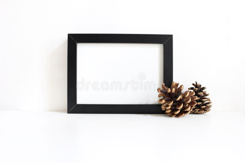 Black blank wooden frame mockup with pine cones lying on the white table. Poster product design. Styled stock feminine photography, home decor. Christmas winter concept. Black blank wooden frame mockup with pine cones lying on the white table. Poster product design. Styled stock feminine photography, home decor. Christmas winter concept.