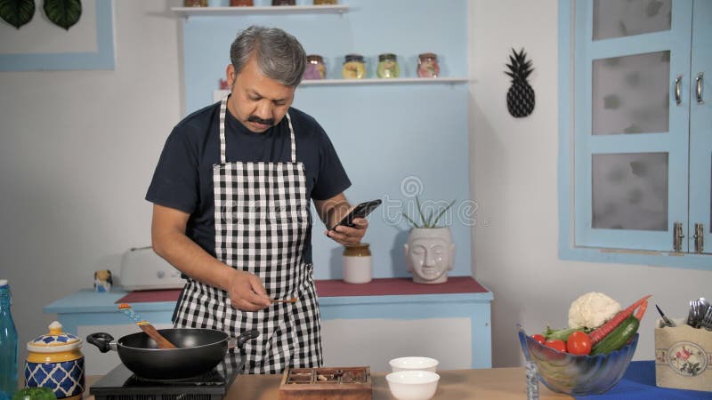 Marido de mediana edad solo en casa comprobando recetas en su móvil para cocinar comidas cocina india