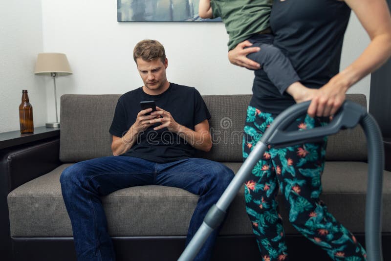 Marito pigro seduto sul divano e al telefono mentre la moglie pulisce casa con un figlio in grembo