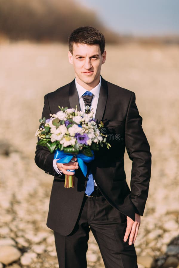 Marié de sourire élégant élégant de jeunes avec le bouquet des fleurs seul marchant sur la plage rocheuse photos stock Marié de sourire élégant élégant de jeunes avec le bouquet des fleurs seul marchant sur la plage rocheuse photos stock