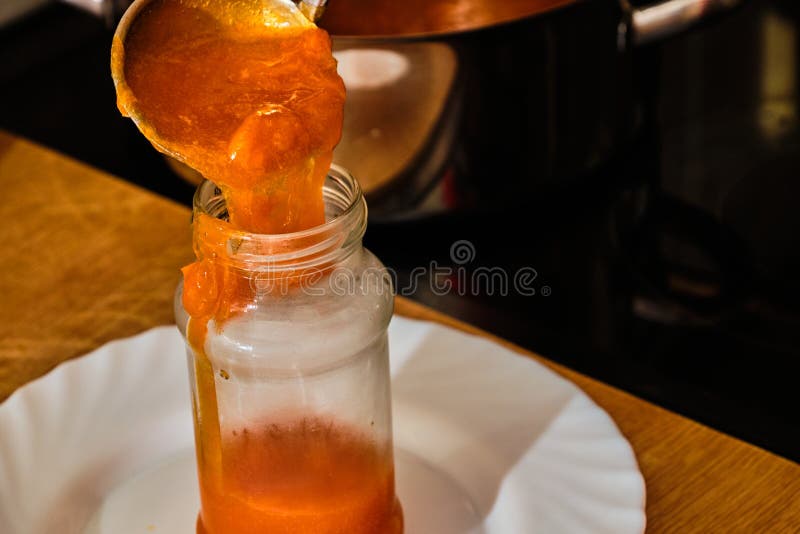 Pouring tasty homemade apricot marmelade in glass with soup ladle on wooden table and view on iron pot on cooker in background. Pouring tasty homemade apricot marmelade in glass with soup ladle on wooden table and view on iron pot on cooker in background