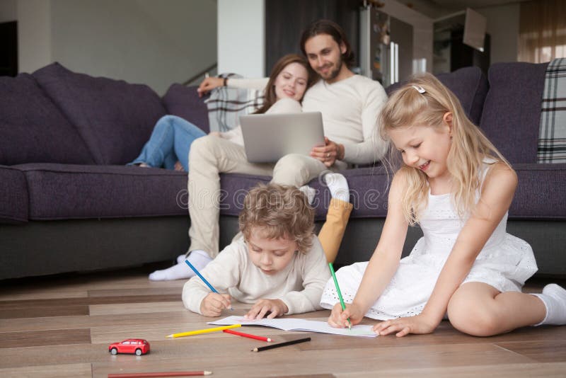 Married relaxed couple with preschool children in living room. Parents sitting on couch using computer, little daughter and son on warm wooden floor drawing. Happy family together at new home concept. Married relaxed couple with preschool children in living room. Parents sitting on couch using computer, little daughter and son on warm wooden floor drawing. Happy family together at new home concept