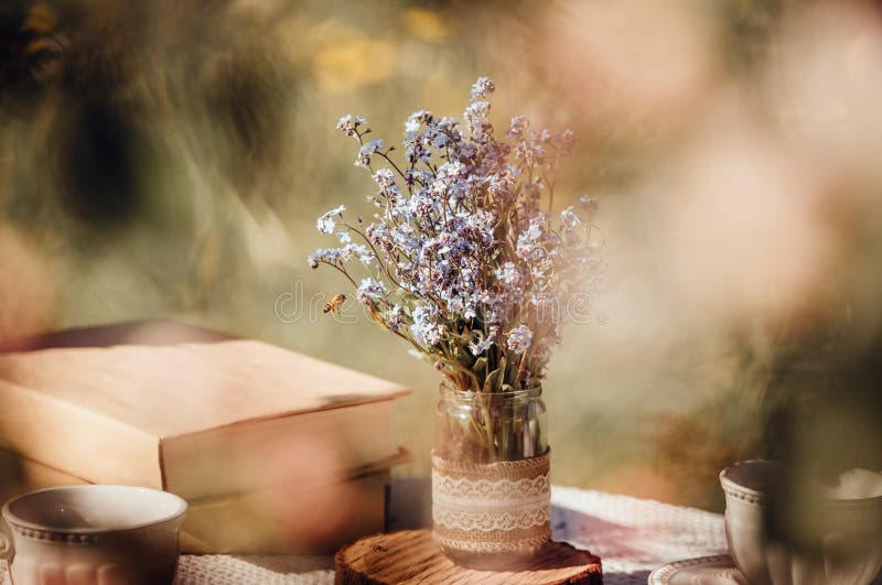 Dream like view through apple blossoms to table, set with tea cups and books. Bouquet of wildflowers Myosotis arvensis forget me nots in handmade jar vase. Dream like view through apple blossoms to table, set with tea cups and books. Bouquet of wildflowers Myosotis arvensis forget me nots in handmade jar vase.