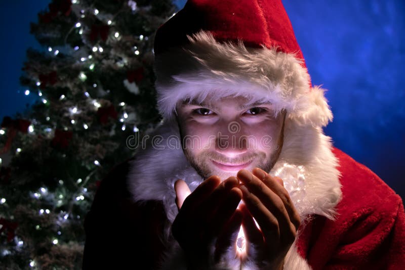 Maschio bello Santa che sorride e che tiene le luci in sue mani e che esamina macchina fotografica con l'albero di Natale nel fon fotografia stock Maschio bello Santa che sorride e che tiene le luci in sue mani e che esamina macchina fotografica con l'albero di Natale nel fon fotografia stock