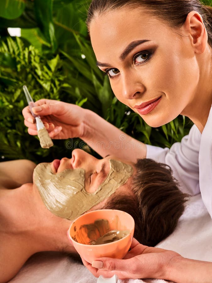 Mud facial mask of men in spa salon. Massage with clay full face. Girl on with therapy room. Man lying spa bed. Beautician with bowl therapeutic procedure green plant background. Mud facial mask of men in spa salon. Massage with clay full face. Girl on with therapy room. Man lying spa bed. Beautician with bowl therapeutic procedure green plant background.