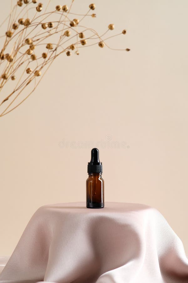 Massage essential oil in amber glass dropper bottle on satin fabric round podium on beige background with dried flax flowers.