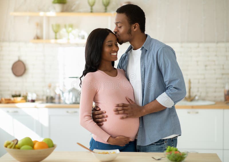 Maternity and healthy eating concept. Caring black husband kissing his pregnant wife in kitchen while cooking breakfast