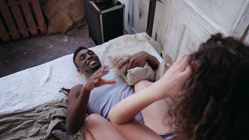 Matin heureux des couples multiraciaux Femme dans des pyjamas se reposant sur l'homme, combat par des oreillers Fille et garçon s banque de vidéos
