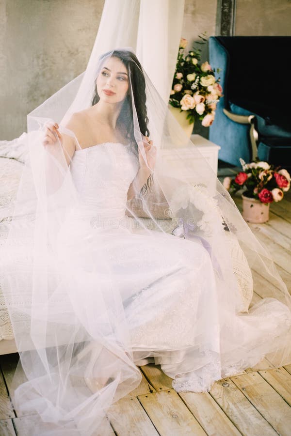 Mattina di una sposa caucasica attraente sexy Sta sorridendo, guardando alla finestra e sta toccando il velo, mentre si sedeva fotografie stock Mattina di una sposa caucasica attraente sexy Sta sorridendo, guardando alla finestra e sta toccando il velo, mentre si sedeva fotografie stock