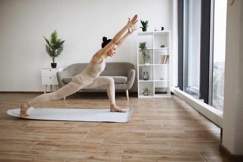 Mature brunette practising Warrior I Halfway Fold during yoga exercise at home. Caucasian woman spread her legs wide and pulls one knee forward, straightening arms up above the head.