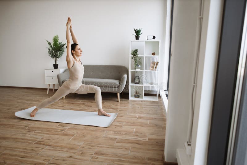 Mature brunette practising Warrior One Pose during yoga exercise at home. Caucasian woman spread her legs wide and pulls one knee forward, straightening arms up above the head.