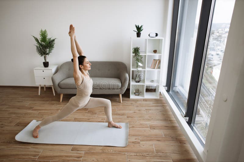 Mature brunette practising Warrior One Pose during yoga exercise at home. Caucasian woman spread her legs wide and pulls one knee forward, straightening arms up above the head.