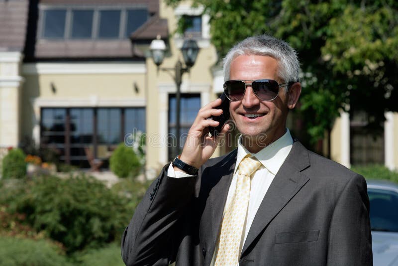 Mature handsome businessman standing before house using mobile phone smiling. Mature handsome businessman standing before house using mobile phone smiling