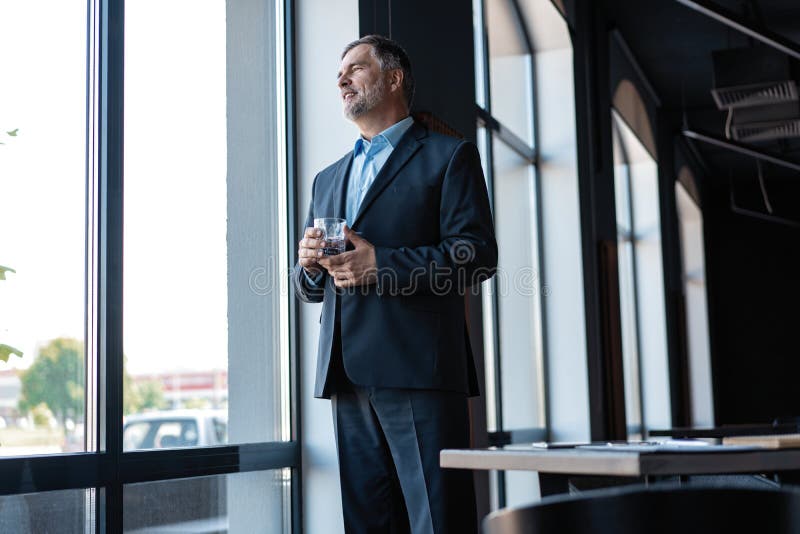 Mature businessman drinking water and looking out of a window at the city from an office building.