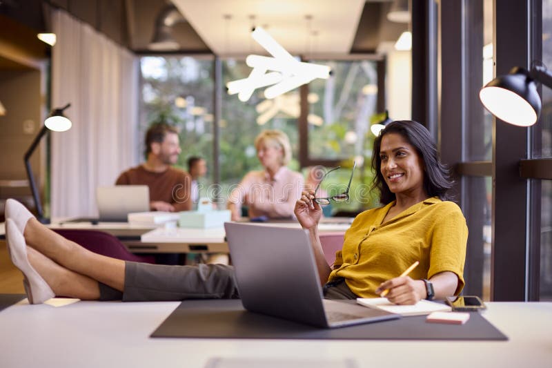 Mature Businesswoman With Feet On Desk Working On Laptop In Office