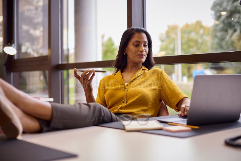 Mature Businesswoman With Feet On Desk Working On Laptop In Office Talking Into Mic Of Mobile Phone