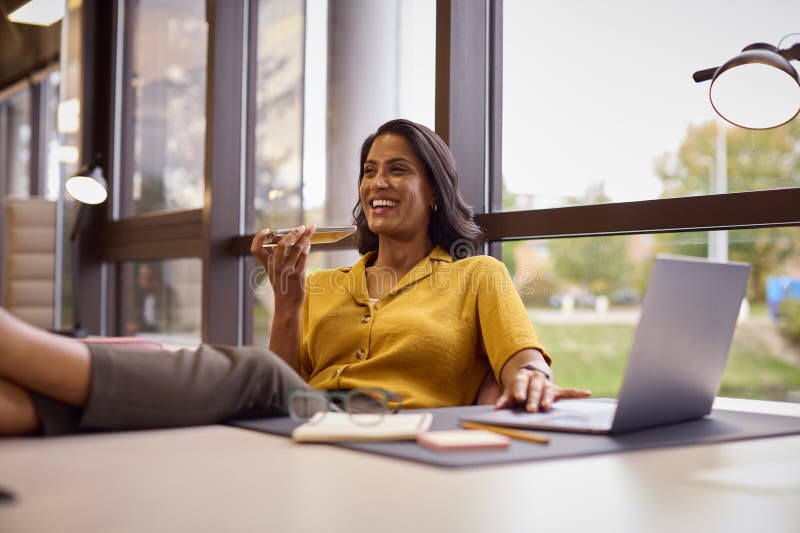 Mature Businesswoman With Feet On Desk Working On Laptop In Office Talking Into Mic Of Mobile Phone