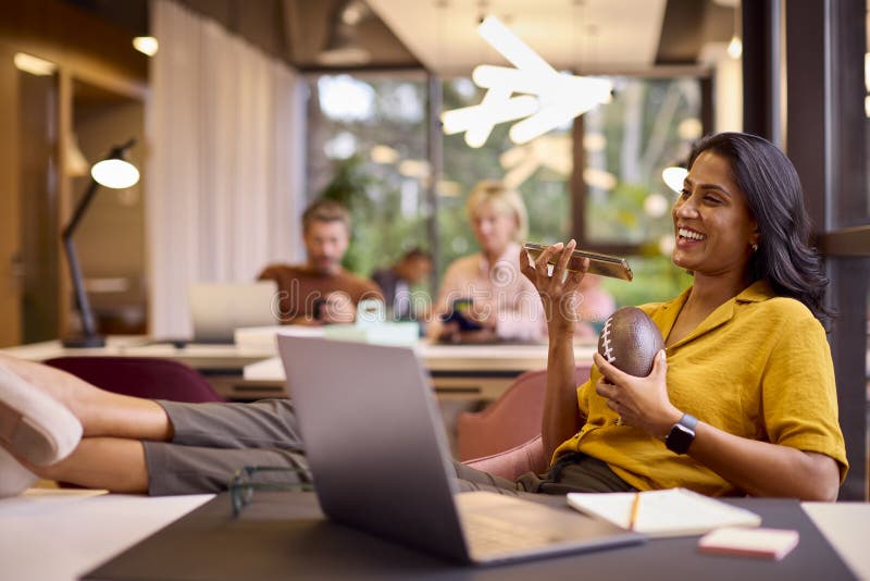 Mature Businesswoman With Feet On Desk Working On Laptop In Office Talking Into Mic Of Mobile Phone