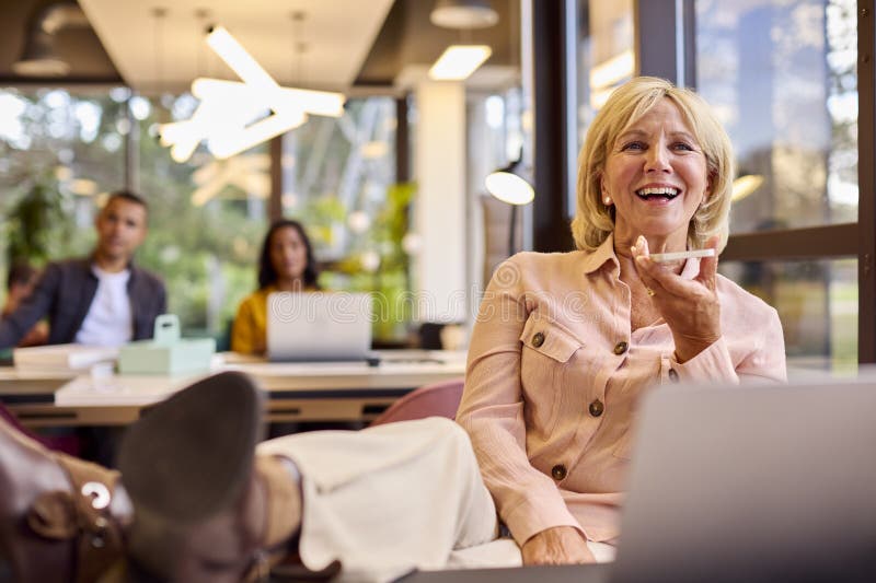 Mature Businesswoman With Feet On Desk Working On Laptop In Office Talking Into Mic Of Mobile Phone