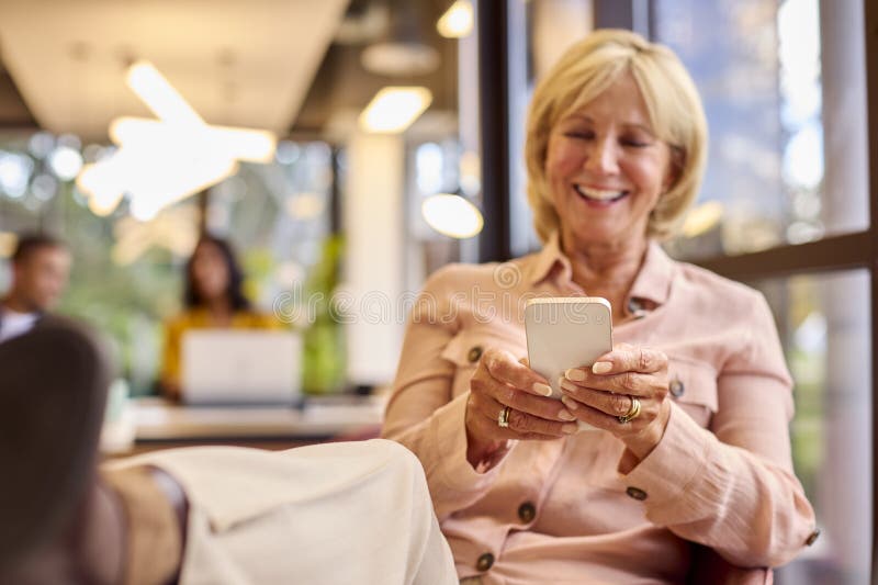 Mature Businesswoman With Feet On Desk Working In Office Checking Messages On Mobile Phone