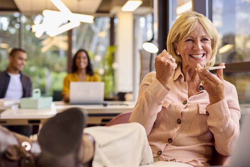 Mature Businesswoman With Feet On Desk Working In Office Talking Into Mic Of Mobile Phone