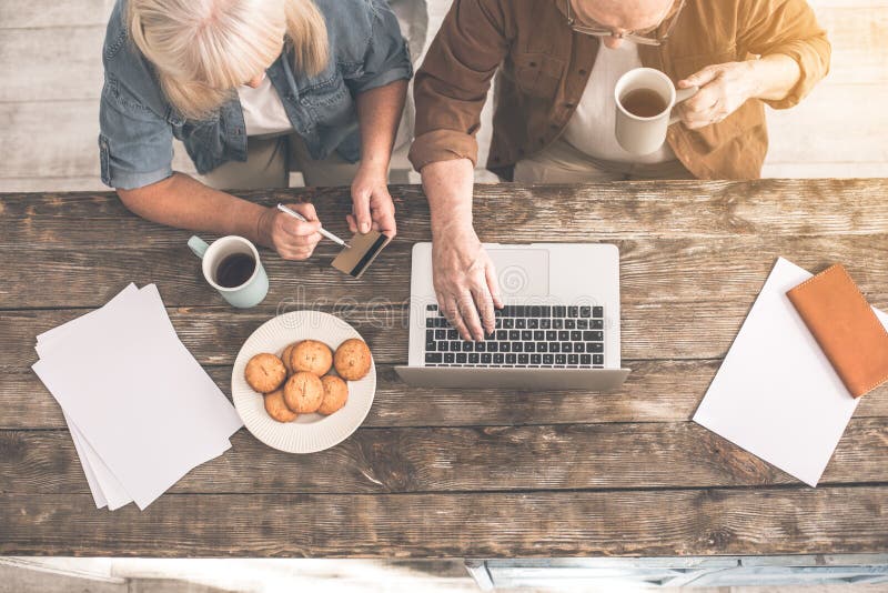 Top view of serious old men typing on modern computer while drinking tea at home. Woman is sitting near him and holding credit card. Online shopping concept. Top view of serious old men typing on modern computer while drinking tea at home. Woman is sitting near him and holding credit card. Online shopping concept