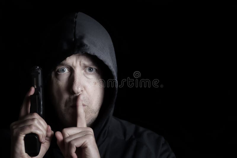 Closeup front view of mature man, looking forward and wearing hood, with weapon and index finger signaling to be silent on dark background. Closeup front view of mature man, looking forward and wearing hood, with weapon and index finger signaling to be silent on dark background