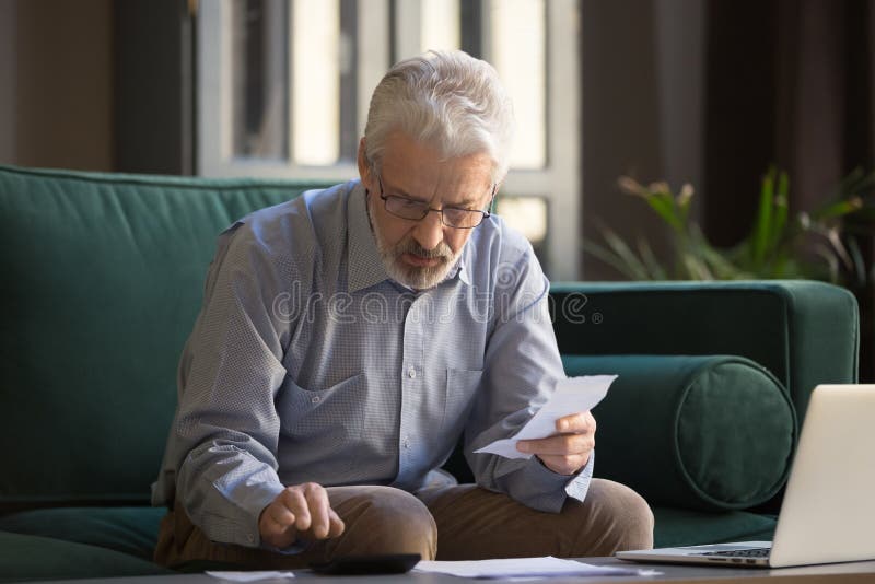 Sixty years old retiree man sit on couch holding receipt paper calculates on calculator monthly expenses, elderly male managing budget, planning finances, analyzes charges, check utility bills concept. Sixty years old retiree man sit on couch holding receipt paper calculates on calculator monthly expenses, elderly male managing budget, planning finances, analyzes charges, check utility bills concept