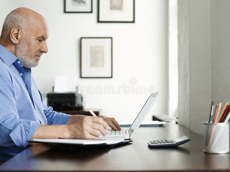 Mature Man Using Laptop And Writing In Notepad
