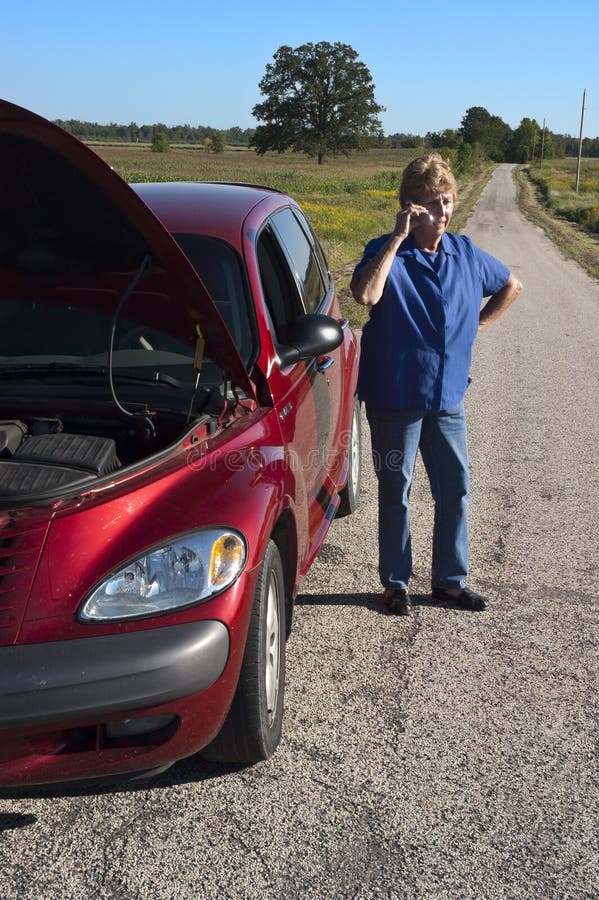 Mature senior female woman with a car broke down and in trouble. The hood is popped up as a signal of distress but being stuck and stranded on a lonely highway means help is not coming anytime soon. It's always extra stress when the auto breaks down and needs repairs. Here we see the lady using her cell phone to call for help. Mature senior female woman with a car broke down and in trouble. The hood is popped up as a signal of distress but being stuck and stranded on a lonely highway means help is not coming anytime soon. It's always extra stress when the auto breaks down and needs repairs. Here we see the lady using her cell phone to call for help.